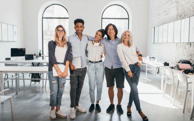 Full-length portrait of shy blonde girl in white sneakers holding laptop after seminar and stands beside african friend. Excited international students posing together after lecture in spacious hall.